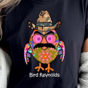 May include: Black t-shirt featuring a colorful owl design wearing a cowboy hat, glasses, and a mustache. The text "Bird Reynolds" is printed below the owl. The word "black" is printed at the bottom.
