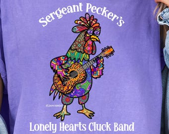 May include: A violet t-shirt with a vibrant illustration of a rooster playing a guitar. The text on the shirt says "Sergeant Pecker's Lonely Hearts Cluck Band". The shirt is a relaxed fit.