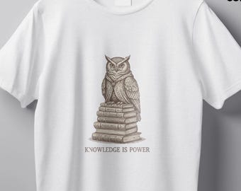 May include: White t-shirt with a sepia-toned illustration of an owl perched atop a stack of books. The text "KNOWLEDGE IS POWER" is printed below the image. The shirt is hanging on a wooden hanger.