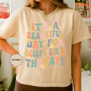 Può includere: T-shirt beige con la scritta "IT'S A BEAUTIFUL DAY FOR OCCUPATIONAL THERAPY" in lettere colorate in stile retrò. Il design include fiori e stelle. La maglietta ha maniche corte e girocollo.