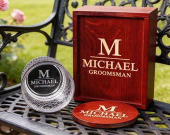 May include: A personalized groomsman gift set featuring a wooden box, a coaster, and a crystal glass. The box and coaster are a dark red color with the letter "M" and the name "MICHAEL GROOMSMAN" engraved in a light color. The glass has a diamond pattern.