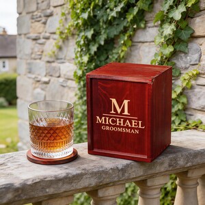 Puede incluir: Una caja de madera de color rojo oscuro con el nombre "MICHAEL GROOMSMAN" y una "M" dorada. Un vaso de líquido ámbar descansa sobre un posavasos de madera. La caja y el posavasos están sobre una superficie de piedra.