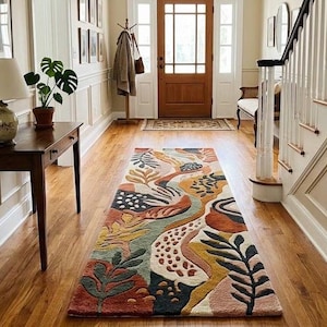 May include: A long rectangular runner rug featuring an abstract botanical pattern with organic shapes and leaves in shades of terracotta, mustard yellow, sage green, cream, and dark navy blue, laid out on a hardwood floor in a residential hallway.