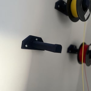 May include: Black 3D-printed filament spool holder mounted on a white wall. The holder is designed to hold a spool of filament, with a black arm extending outward. Two spools of filament are visible, one yellow and one red.