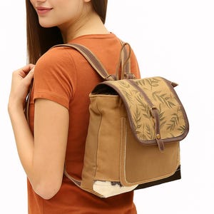 May include: A tan canvas backpack with a flap closure, featuring a leaf pattern. The backpack has brown leather straps and trim. The bag is worn over the shoulders.
