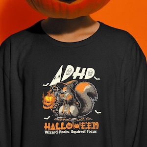 May include: Black sweatshirt with a Halloween-themed graphic. The design features a squirrel dressed as a wizard holding a jack-o'-lantern. Text includes "ADHD" and "HALLOWEEN" with the phrase "Wizard Brain. Squirrel focus."