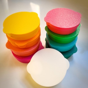 May include: Stacks of colorful, round, plastic food storage container lids in yellow, orange, pink, red, green, and teal. A white, round container with two side handles sits in front of the stacks.