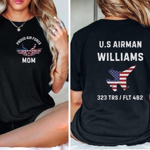 May include: Black t-shirts with patriotic designs. One shirt features "PROUD AIR FORCE MOM" with an American flag graphic. The other displays "U.S AIRMAN WILLIAMS" with an American flag jet graphic and "323 TRS / FLT 482".