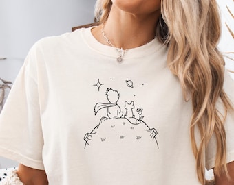 May include: Cream-colored t-shirt featuring a black line drawing of a figure with a scarf, a fox, and a flower on a small planet, with stars and a planet in the background. The shirt has a relaxed fit.
