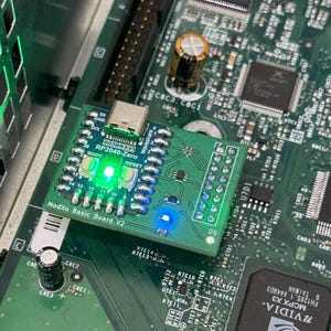 May include: A green ModXo Basic Board V2 circuit board with a Waveshare RP2040-Zero module, featuring multiple LED lights. The board is mounted on a larger green circuit board with various electronic components.