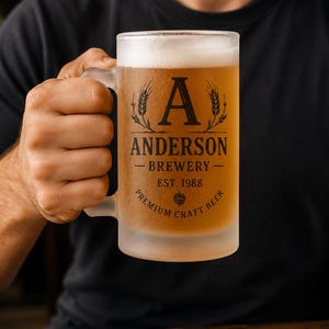 Puede incluir: Una jarra de cerveza de vidrio esmerilado llena de cerveza dorada y espuma. La jarra presenta un diseño impreso en negro con una letra A grande, el texto ANDERSON BREWERY, EST. 1988 y las palabras PREMIUM CRAFT BEER alrededor de un icono de lúpulo.