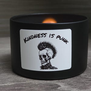 Könnte beinhalten: Eine schwarze Kerze in einem runden Behälter mit einer brennenden orangefarbenen Flamme. Ein weißes Etikett auf der Vorderseite zeigt einen Totenkopf mit Irokesenschnitt und dem Text "KINDNESS IS PUNK". Die Kerze steht auf einer Holzoberfläche.