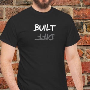 May include: Black t-shirt with the words "BUILT" above "DIFF" in white and gray lettering. The word "DIFF" is a mirrored reflection of the word "BUILT". The shirt is made of a soft material.
