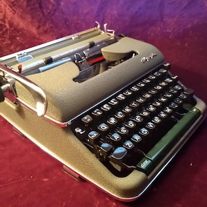 May include: A vintage olive green typewriter with black keys and a silver carriage return lever. The typewriter has a green knob on the left side and the word "Royal" is visible on the top. The keys are arranged in a QWERTY layout.