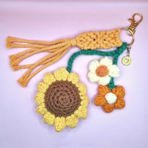 May include: A handmade crochet keychain featuring a sunflower and two smaller flowers in yellow, brown, and orange. The keychain includes a gold-colored clasp and a tassel. The background is a light pink.