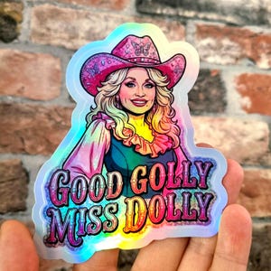 Puede incluir: Una pegatina holográfica con una ilustración estilizada de una persona rubia con un sombrero de vaquero rosa con detalles de mariposas. El texto "GOOD GOLLY MISS DOLLY" está impreso en letras negritas, coloridas y degradadas en la parte inferior.
