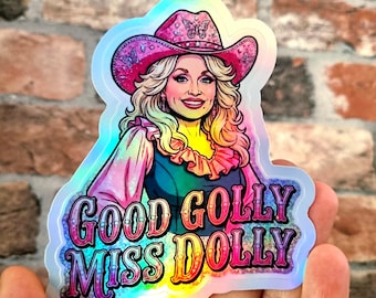 May include: A holographic sticker featuring a stylized illustration of a blonde person wearing a pink cowboy hat with butterfly accents. The text "GOOD GOLLY MISS DOLLY" is printed in bold, colorful, gradient lettering at the bottom of the sticker.