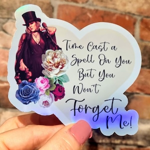Puede incluir: Una pegatina en forma de corazón con una mujer con sombrero de copa y micrófono, rodeada de flores. La pegatina tiene el texto "Time Cast a Spell On You But You Won't Forget Me!" en escritura negra. La pegatina tiene un acabado holográfico.