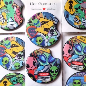 May include: Round car coasters featuring a colorful collage of alien, UFO, and cow abduction graphics. The designs include yellow warning signs, green extraterrestrial faces, and red danger labels with the text Restricted Area.