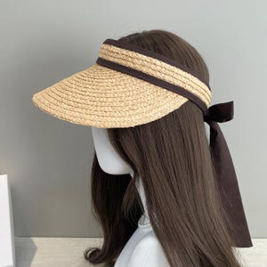 May include: A straw visor with a wide brim and a dark brown band and ribbon. The visor is made of woven straw and has a curved shape to provide shade. The dark brown ribbon is tied in a bow at the back.