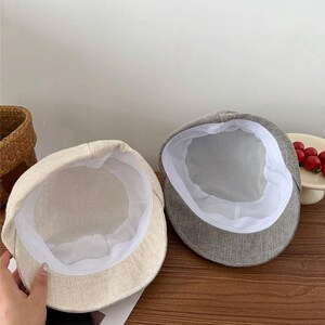 Op de afbeelding: Twee petten in newsboy-stijl. E&eacute;n is lichtbeige van kleur en de andere grijs. Beide petten hebben een witte voering en een ronde kroon. De petten zijn gemaakt van een textielstof.