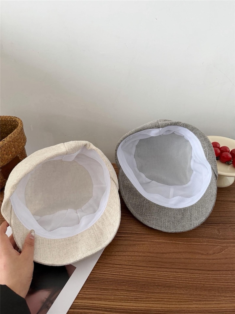 Op de afbeelding: Twee petten in newsboy-stijl. E&eacute;n is lichtbeige van kleur en de andere grijs. Beide petten hebben een witte voering en een ronde kroon. De petten zijn gemaakt van een textielstof.