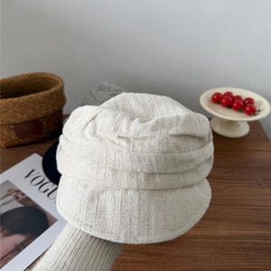 Op de afbeelding: Een lichtbeige newsboy-pet van een textuurstof. De pet heeft een afgeronde kroon en een kleine, stijve klep. Het woord "VOGUE" is gedeeltelijk zichtbaar op een tijdschrift op de achtergrond. Een klein mandje en een bordje met rode bessen zijn ook op de achtergrond.