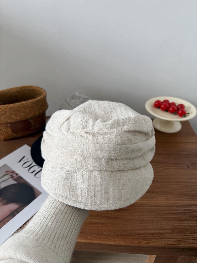 Op de afbeelding: Een lichtbeige newsboy-pet van een textuurstof. De pet heeft een afgeronde kroon en een kleine, stijve klep. Het woord "VOGUE" is gedeeltelijk zichtbaar op een tijdschrift op de achtergrond. Een klein mandje en een bordje met rode bessen zijn ook op de achtergrond.
