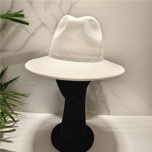 May include: A solid off-white fedora hat with a structured crown and a wide, flat brim. The hat features a smooth, felt-like texture and is displayed on a black mannequin head stand, showcasing its clean lines and neutral color palette.