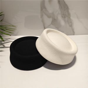 May include: Two pillbox hats, one in solid black and one in solid white, featuring a smooth, felted texture with a flat top and a structured, cylindrical side profile. These hats are positioned side-by-side, showcasing their simple, circular design.