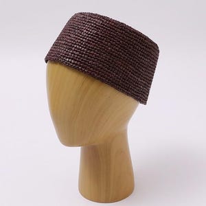 May include: A dark brown pillbox hat crafted from a woven, textured material. The hat features a structured, cylindrical shape with a flat top and a consistent, tight weave pattern throughout the entire exterior surface of the headwear piece.