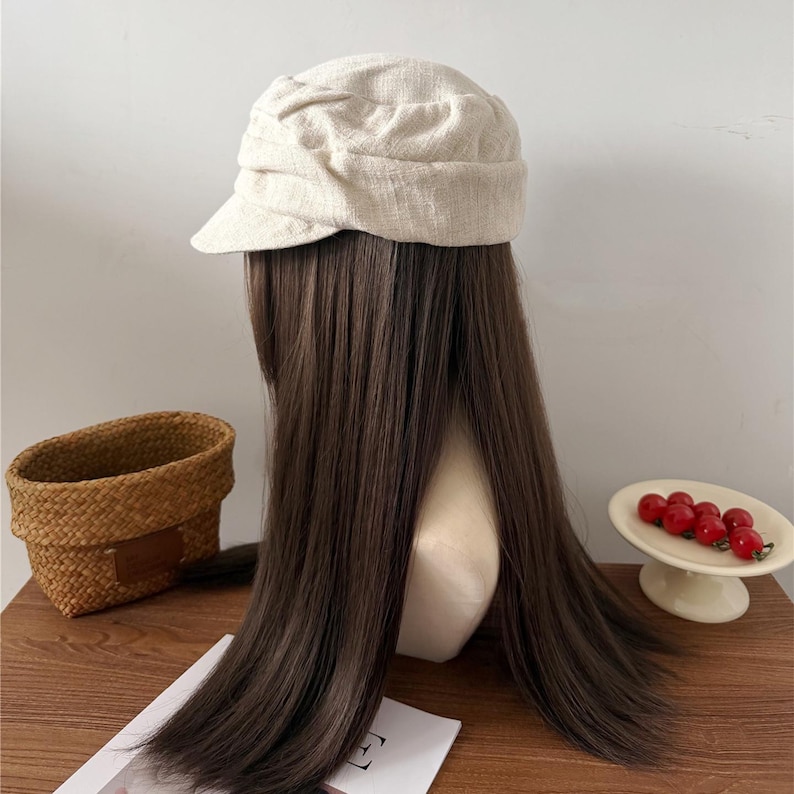 Op de afbeelding: Een beige linnen baret zit op een mannequinhoofd met lang, donkerbruin haar. De baret heeft een afgeronde kroon en een kleine, stijve rand. Een geweven mand en een klein schaaltje met rode kersen staan op de achtergrond.
