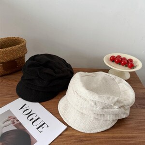 Op de afbeelding: Twee petten in newsboy-stijl, &eacute;&eacute;n zwarte en &eacute;&eacute;n gebroken witte, liggen op een houten oppervlak. De gebroken witte pet is gemaakt van een textuurstof. Een exemplaar van het tijdschrift Vogue is zichtbaar. Een klein mandje en een schaal met rode bessen staan op de achtergrond.
