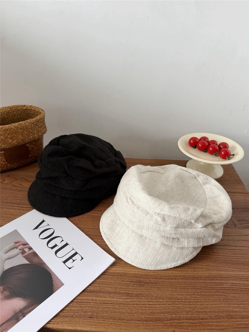 Op de afbeelding: Twee petten in newsboy-stijl, &eacute;&eacute;n zwarte en &eacute;&eacute;n gebroken witte, liggen op een houten oppervlak. De gebroken witte pet is gemaakt van een textuurstof. Een exemplaar van het tijdschrift Vogue is zichtbaar. Een klein mandje en een schaal met rode bessen staan op de achtergrond.