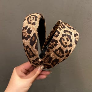 Peut inclure: Un serre-tête large et rembourré arborant un motif léopard dans des tons beige et marron. L'accessoire présente un profil arrondi et surélevé avec une doublure intérieure noire. La surface en tissu offre un aspect texturé et doux.