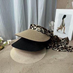 May include: Three sun visors in a stack. The top visor is tan with a leopard print band and a matching scarf. The middle visor is black, and the bottom visor is a light beige color. The visors are made of woven material.