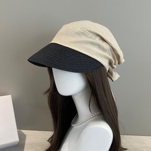 May include: A hat featuring a dark charcoal grey woven straw brim and a light beige fabric crown. The fabric crown is soft and unstructured, draping slightly at the back, and includes fabric ties that allow for an adjustable fit around the head.