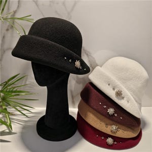 May include: A black felt cloche hat on a display stand, with a stack of four additional cloche hats in white, brown, tan, and burgundy. Each hat features a wide brim and is decorated with a cluster of small, clear, sparkling crystal accents.