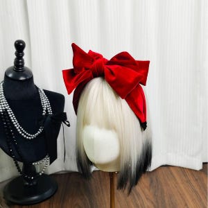 May include: A red velvet bow headband is displayed on a mannequin head with white and black ombre hair. A black bust with pearl necklaces and a black bow is to the left. The background is a white curtain.