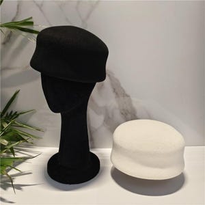 May include: A black felt pillbox hat displayed on a black mannequin head and a separate white felt pillbox hat resting on a surface. Both hats feature a structured, rounded crown with a flat top and a short, slightly flared brim design.