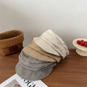 Op de afbeelding: Een stapel petten in verschillende neutrale kleuren, waaronder grijs, beige en bruin. De petten zijn gemaakt van een textuurstof en liggen op een houten oppervlak. Een tijdschrift met het woord "VOGUE" is zichtbaar.
