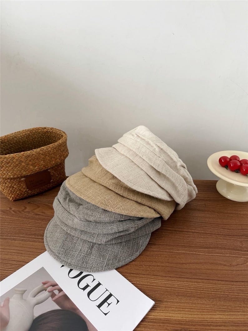 Op de afbeelding: Een stapel petten in verschillende neutrale kleuren, waaronder grijs, beige en bruin. De petten zijn gemaakt van een textuurstof en liggen op een houten oppervlak. Een tijdschrift met het woord "VOGUE" is zichtbaar.