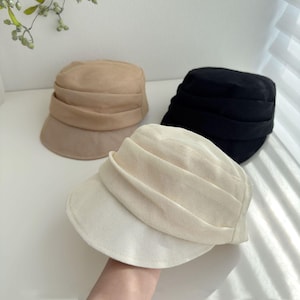 Op de afbeelding: Drie stijlvolle hoeden in beige, zwart en gebroken wit. Elke hoed heeft een korte rand en een geplooide kroon. De hoeden zijn gemaakt van een zachte, getextureerde stof, perfect voor elke outfit.