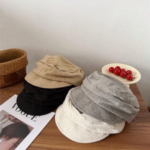 Op de afbeelding: Een verzameling baretmutsen in verschillende kleuren, waaronder beige, zwart, grijs en wit, is op een houten oppervlak gestapeld. Een exemplaar van het tijdschrift Vogue en een kleine mand zijn ook zichtbaar. Een klein bordje met rode kerstomaatjes is ook aanwezig.