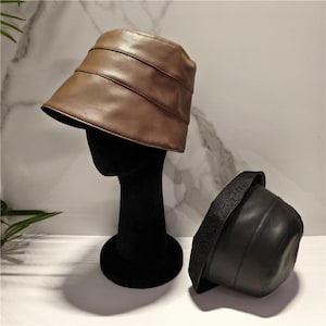 May include: A brown bucket hat with horizontal stitched paneling sits on a black mannequin head, while a second black bucket hat with a textured brim is positioned next to it. Both hats feature a smooth finish and a rounded crown design.