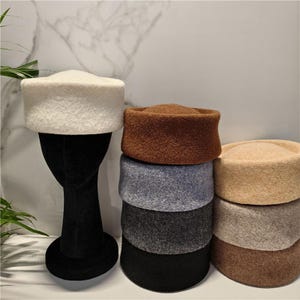 May include: A collection of pillbox hats in various colors including white, brown, light beige, grey, dark grey, and black. The hats feature a textured, felt-like material with a flat top and a structured, cylindrical shape, displayed on a black stand.