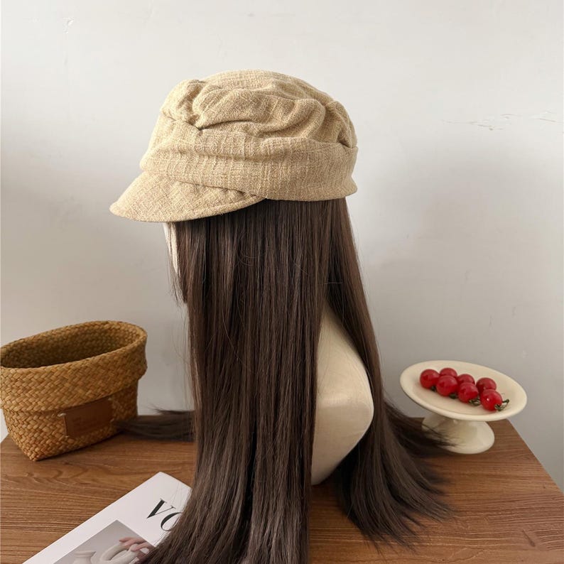 Op de afbeelding: Een beige newsboy-pet met een korte klep. De pet is gemaakt van een textuurstof en staat op een mannequinhoofd met lang, donkerbruin haar. Op de achtergrond staan een gevlochten mand en een klein schaaltje met rood fruit.