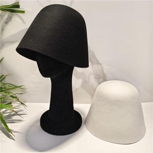 Puede incluir: Un sombrero de pescador de fieltro negro liso sobre una cabeza de maniquí negra, acompañado de un sombrero de pescador de fieltro blanco liso colocado al lado. Ambos sombreros presentan una copa redondeada lisa y un ala corta inclinada.