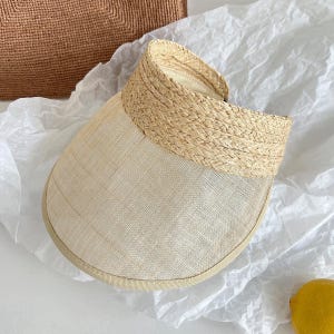 May include: A beige sun visor featuring a wide, semi-transparent brim with a fine woven texture and a structured, braided straw headband. The visor has a clean, neutral color palette and a curved design that provides shade while maintaining a lightweight look.