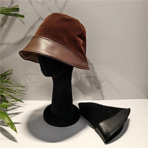May include: A brown bucket hat featuring a soft suede crown and a smooth leather brim. Beside it lies a second bucket hat in black, also constructed with a suede crown and a leather brim. Both hats display a structured design with a wide brim.